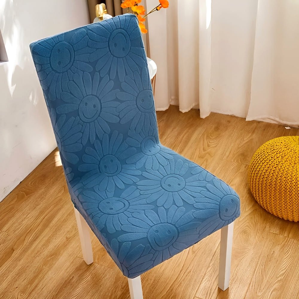 Blossom Aura stretch chair cover in Teal Breeze, featuring a blue floral fabric with a unique smiley face daisy pattern. This elastic fit cover protects furniture and adds stylish home decor, shown on a dining chair.
