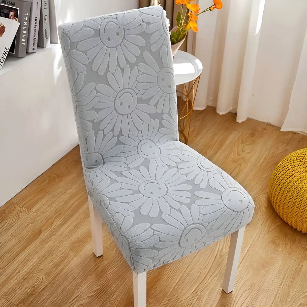 Frost Gray Blossom Aura stretch chair cover featuring a floral daisy pattern with smiling faces on a white-legged chair in a home setting.