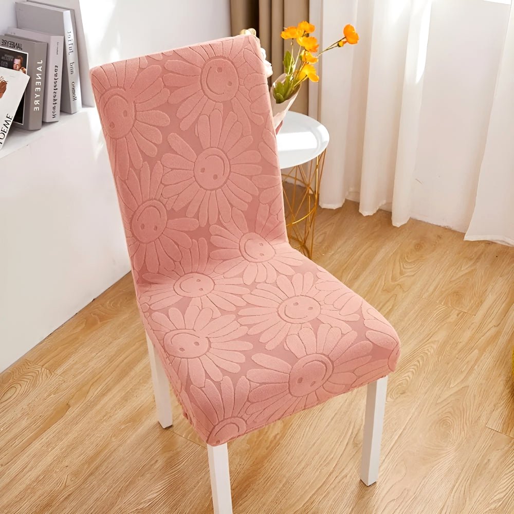 Rosy Blush Blossom Aura stretch chair cover featuring a floral daisy pattern with smiling faces on a white-legged chair in a home setting.