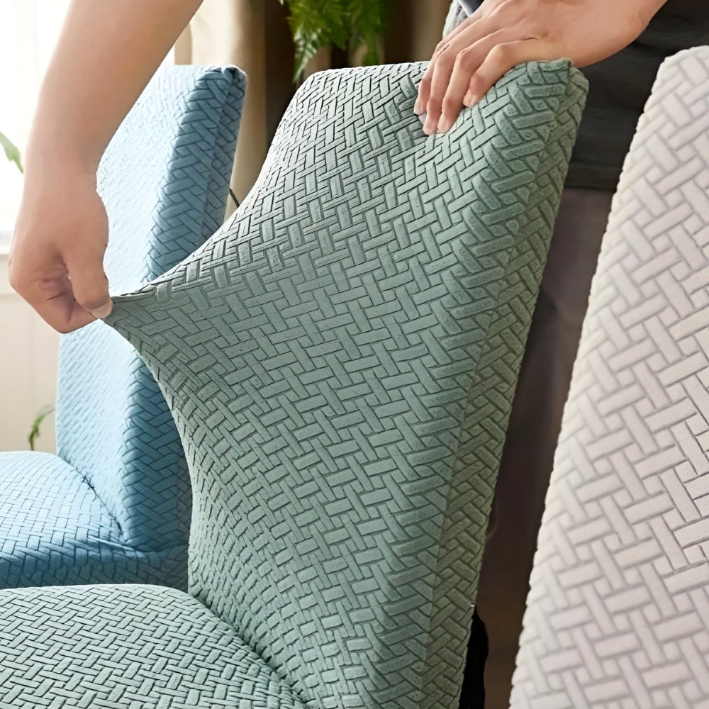 A person's hands stretching a Veluxe Haven Jacquard Velvet Dining Chair Cover in olive green, demonstrating its flexible stretch fit and textured jacquard pattern, with other covered chairs in the background.
