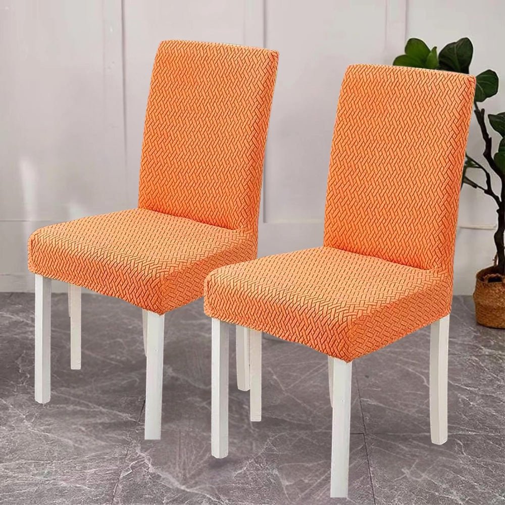 Two dining chairs covered in vibrant tangerine orange jacquard velvet stretch fit covers, showcasing the textured fabric and white chair legs.