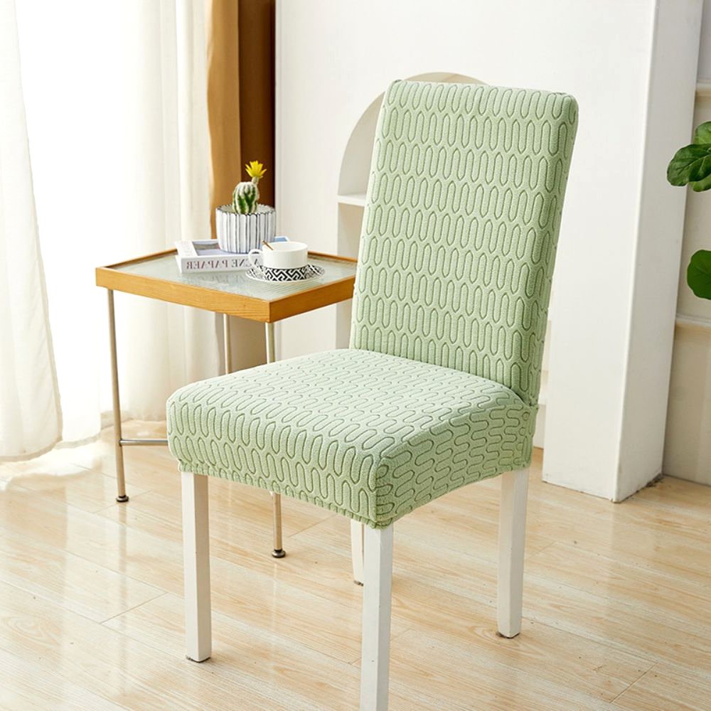 Sage green elastic jacquard chair cover with a chevron maze pattern, shown on a dining chair in a modern room setting with a side table and plant.