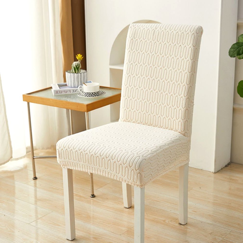 Soft white elastic jacquard chair cover with a chevron maze pattern, shown on a dining chair in a modern room setting with a side table and plant.