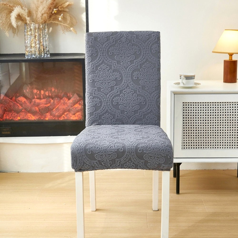 Midnight Slate Velvet Haven elegant jacquard chair seat slipcover on a dining chair in a modern room setting with a side table and fireplace.