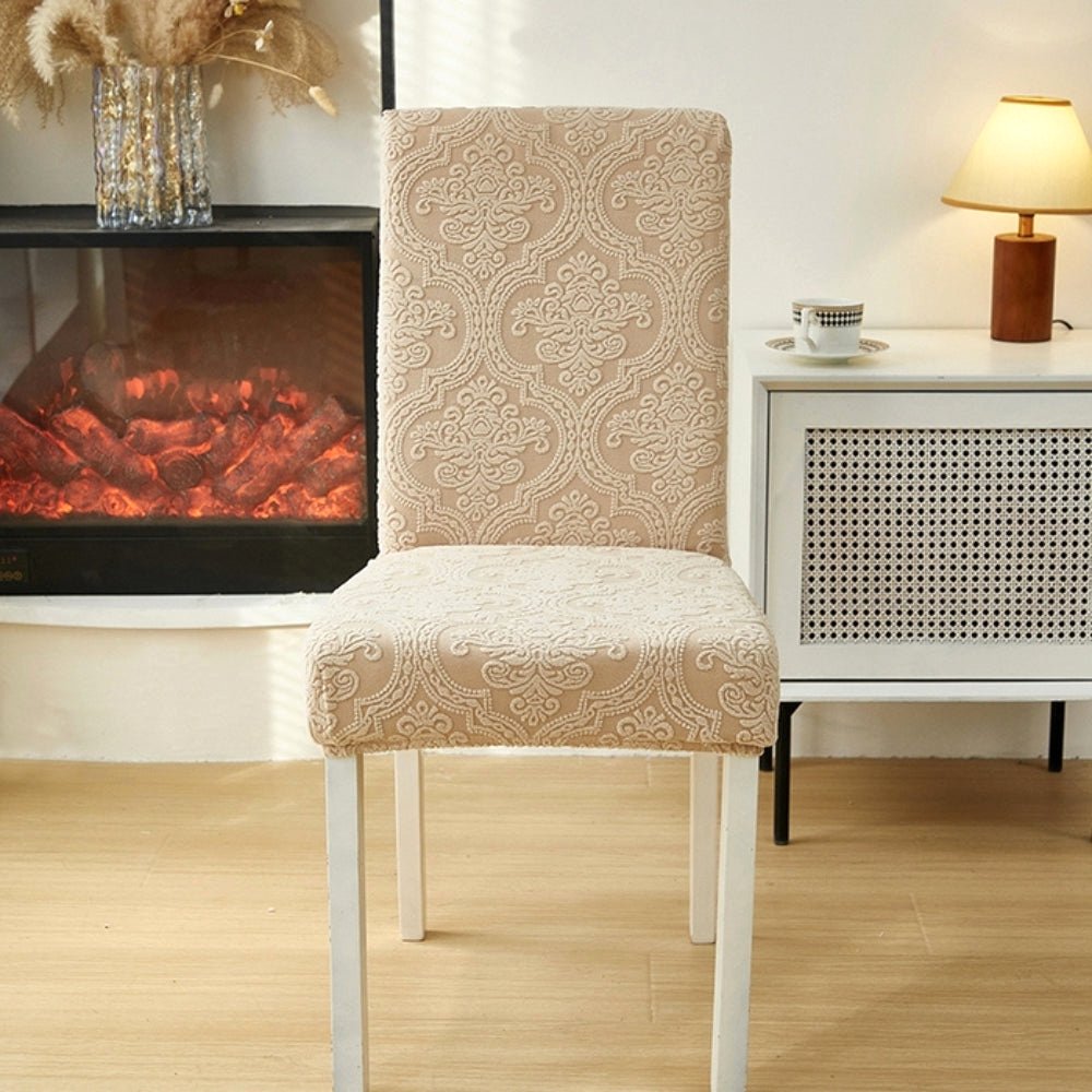 Sand Bloom Velvet Haven elegant jacquard chair seat slipcover on a dining chair in a cozy room setting with a lamp and fireplace.