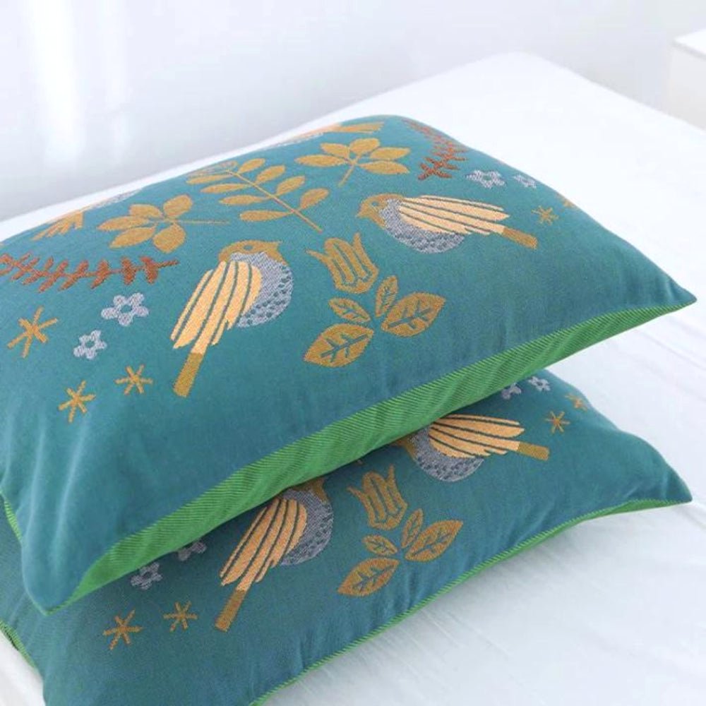 Two Nordic Nest cotton gauze pillowcases with a soft texture, featuring a teal color and a charming bird and leaf pattern, stacked on a bed. These decorative pillows offer a cozy and breathable fabric for home decor.