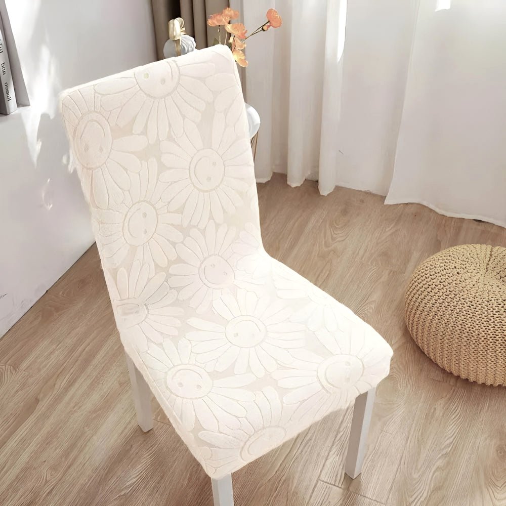 Cream Blossom stretch chair cover in floral fabric with elastic fit, featuring a smiling daisy pattern, on a dining chair in a bright room.