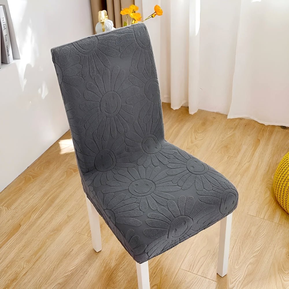 Blossom Aura stretch chair cover in Graphite Bloom, featuring a dark gray floral fabric with a unique smiley face daisy pattern. This elastic fit cover protects furniture and adds stylish home decor, shown on a dining chair.