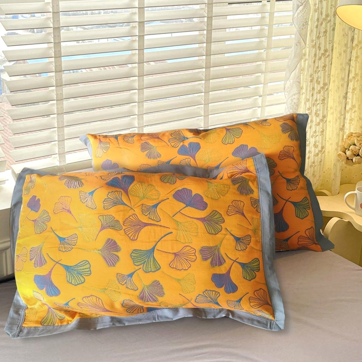A pair of Ginkgo Haven cotton pillowcases, yellow with embroidered blue green ginkgo leaf patterns grey borders, lying side-by-side on a white bed window blinds in background.