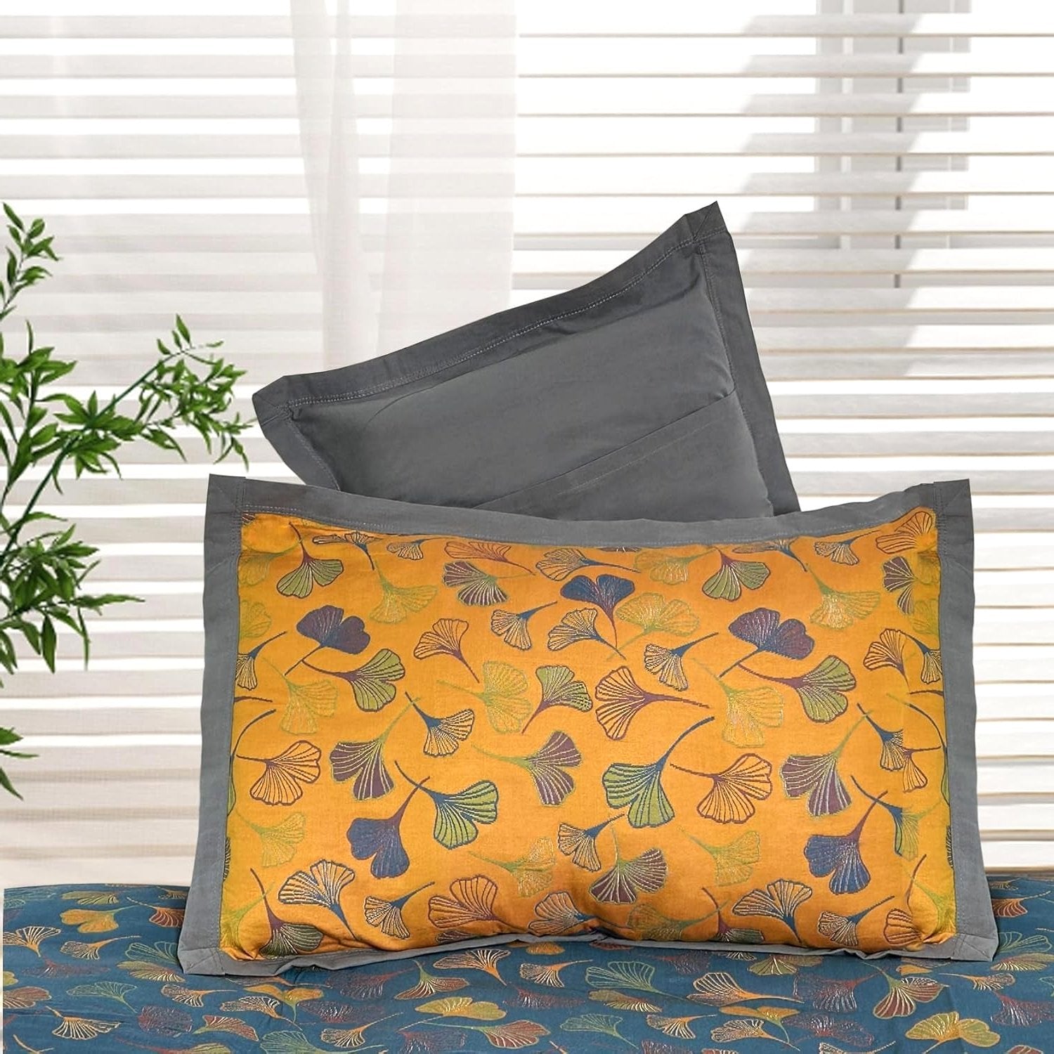 A pair of Ginkgo Haven cotton pillowcases, one yellow with embroidered blue green ginkgo leaf patterns grey border, stacked on a bed plain grey pillowcase underneath. A window blind green plant are in background.