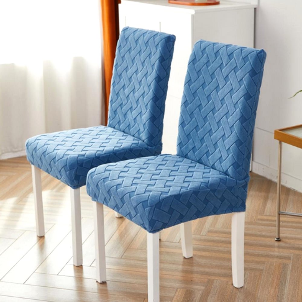 Two dining chairs fitted with a Sky Mist Blue Haven Weave Jacquard Chair Cover, displaying its elegant woven design. The chairs have white legs and are situated in a well-lit room with a light wood floor.