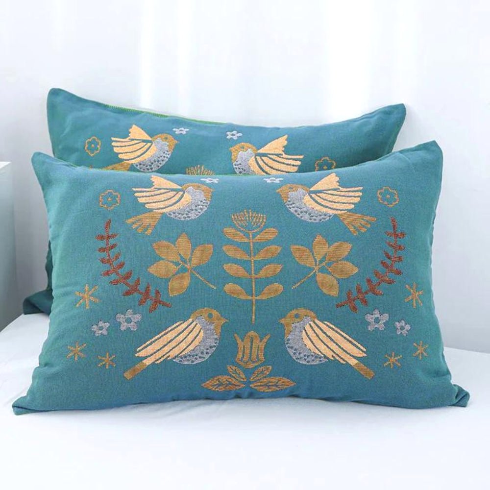 Two Nordic Nest cotton gauze pillowcases in teal, featuring intricate embroidered bird and botanical patterns in gold and brown, displayed on a bed. Showcases the decorative and Nordic style.