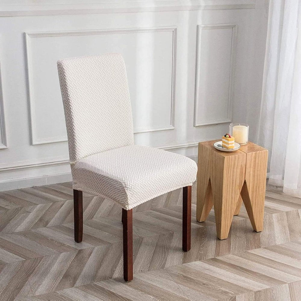 A cream beige jacquard velvet stretch fit dining chair cover on a chair, next to a wooden stool with a dessert, showcasing the textured fabric.