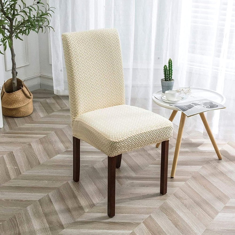 A cream beige jacquard velvet stretch fit dining chair cover on a chair in a dining room setting, featuring a textured pattern and dark wooden legs.