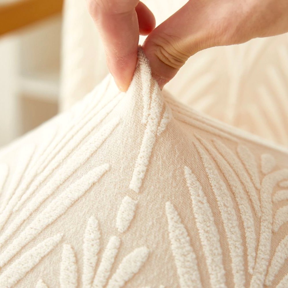 Close-up of a hand stretching the soft white Velvet Haven elastic jacquard chair cover, highlighting its stretch fit and textured palm weave pattern.