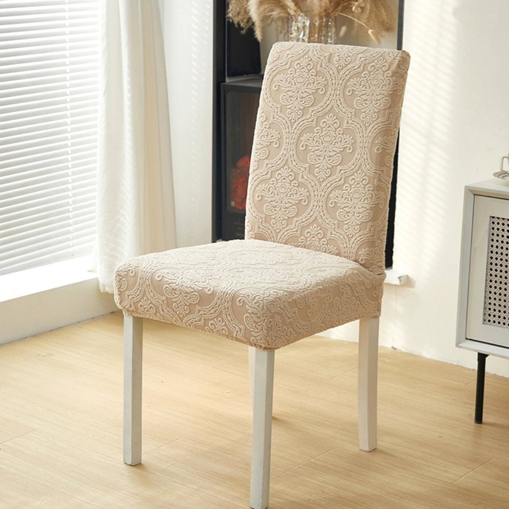 A single dining chair covered in a Sand Bloom elegant jacquard stretch fit slipcover, positioned in front of a window with blinds, highlighting the fabric's texture.