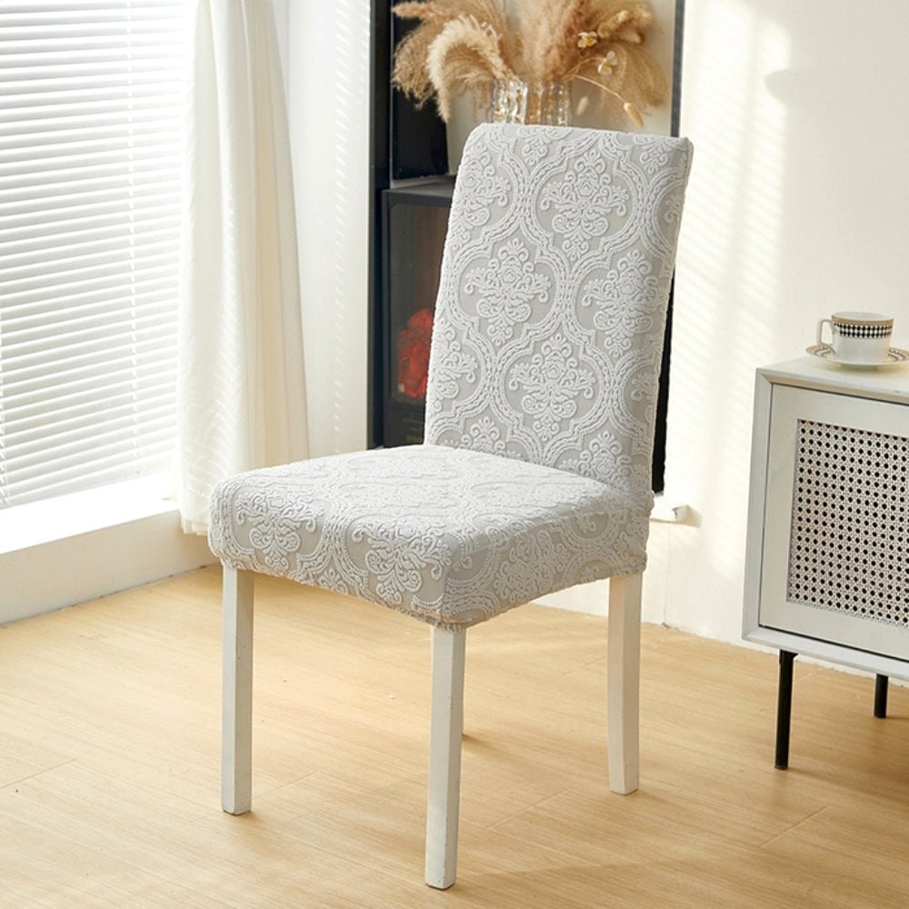 Ash Mist Velvet Haven elegant jacquard chair seat slipcover on a dining chair in a modern room setting with a side table and window.
